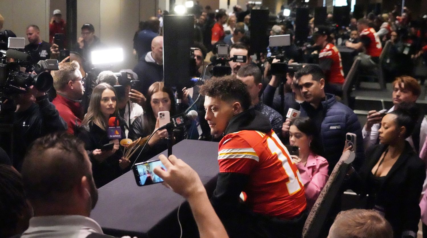 Patrick Mahomes sits at a table with a microphone surrounded by reporters and cameras
