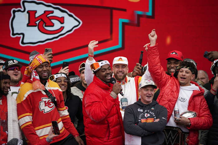 Chris Jones talks into a mic while Travis Kelce, Patrick Mahomes and various other Chiefs players put their hands in the air celebrating at the Super Bowl parade