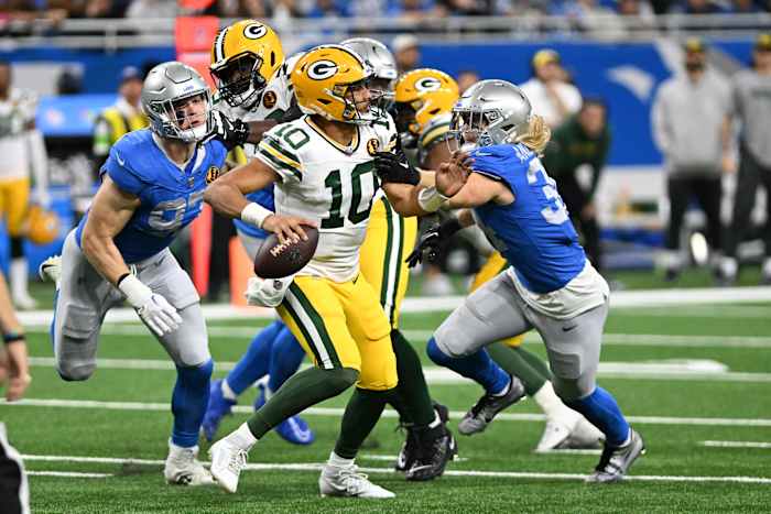 Jordan Love leans back with the ball in one hand to throw while Lions players push Packers players off to reach him