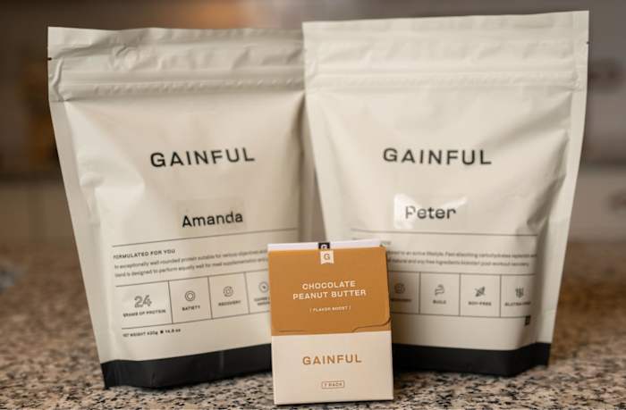 2 Gainful protein powder white bags one labeled "Amanda" and the other labeled "Peter" with a small chocolate peanut butter sample in front sitting on a counter top