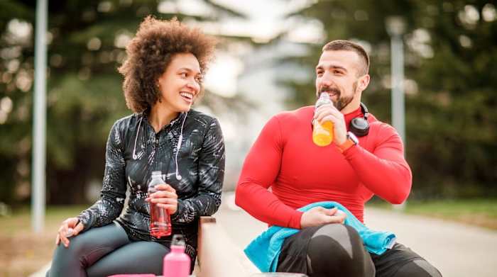 A man and woman sitting on a park bench drinking hydration beverages