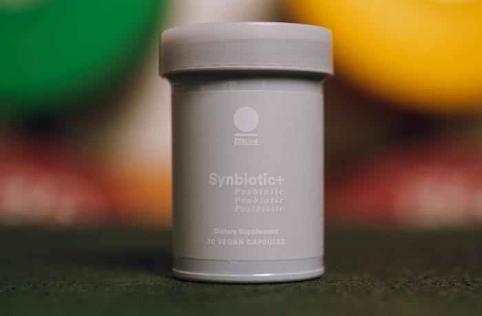 A light blue bottle of Ritual Synbiotic+ probiotic supplement with blurred weight plates in the background