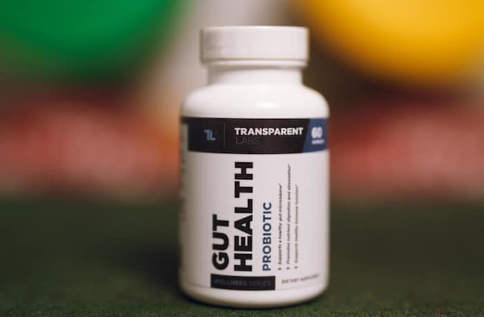 Transparent Labs Gut Health Probiotic white bottle with blue and black writing with blurred weight plates in the background