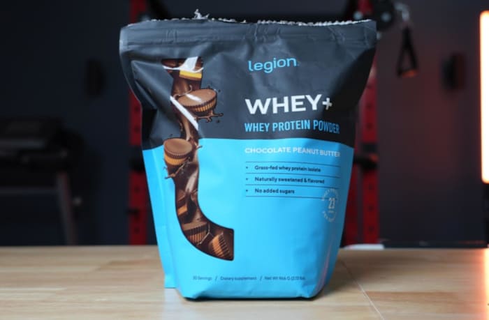 A blue and black bag of Legion Whey+ protein powder in Chocolate Peanut Butter flavor against a home gym backdrop