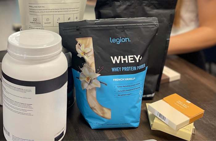 A bag of Legion Whey+ protein powder in French Vanilla flavor on a table with other bags and containers of protein powder.