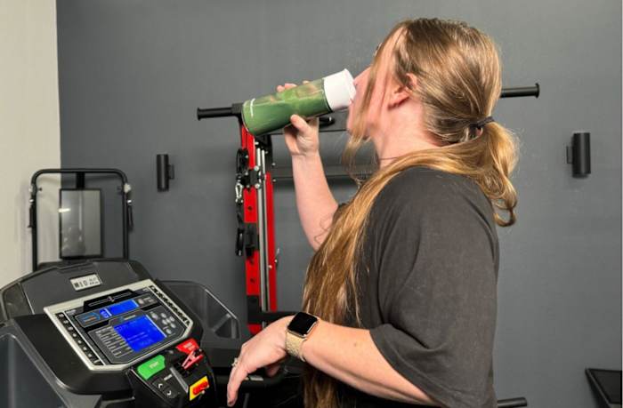 A woman drinking Cymbiotika plant protein from a shaker bottle while walking on a treadmill