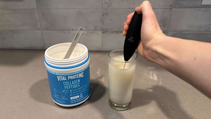 A person using an electric frother to mix unflavored Vital Proteins Collagen Peptides with water in a clear glass