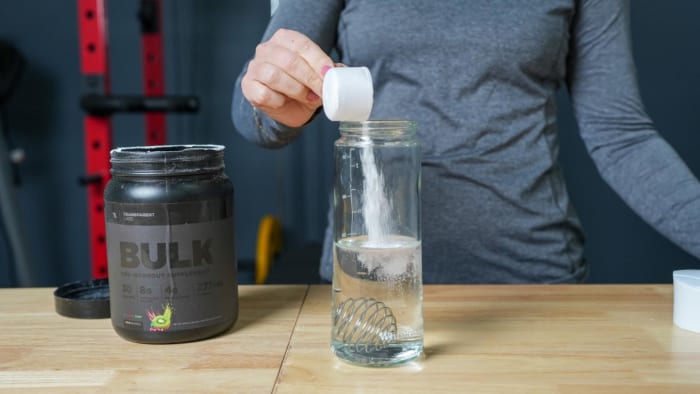 A woman pouring a scoop of Strawberry Kiwi flavor Transparent Labs BULK Black pre-workout in a shaker bottle filled with water