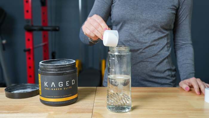 A woman in a gym pouring a scoop of Kaged Pre-Kaged Elite pre-workout powder into a shaker bottle filled with water