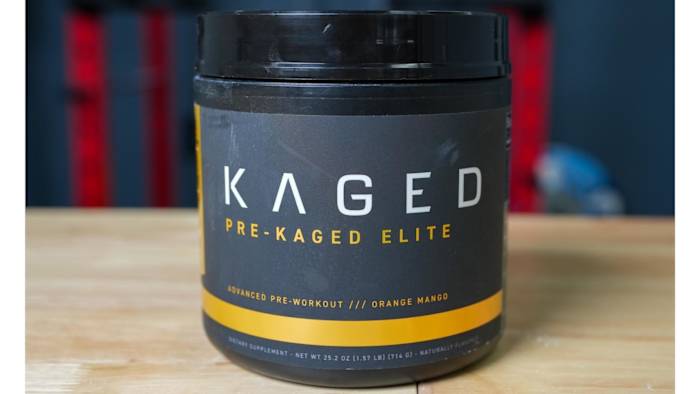 A black and orange tub of Kaged Pre-Kaged Elite pre-workout supplement in Orange Mango flavor on a wooden table in a gym