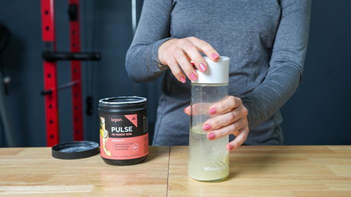 A woman in a gym mixing a serving of Legion Pulse pre-workout in a shaker bottle filled with water