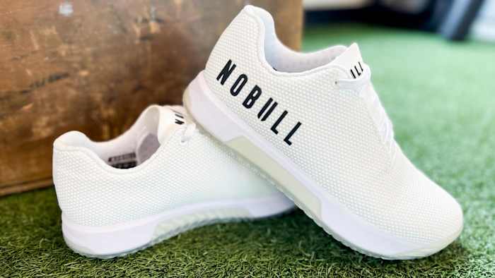 Close up photo of white Nobull Trainer+ shoes sitting on turf.