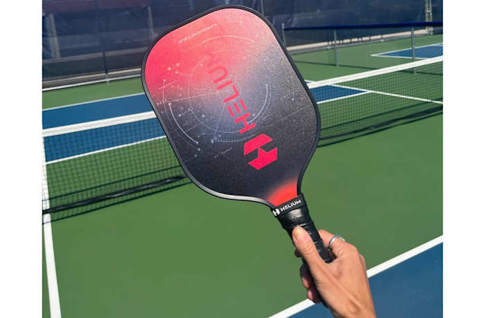 Hand holding a red and black Helium paddle with a pickleball court background.