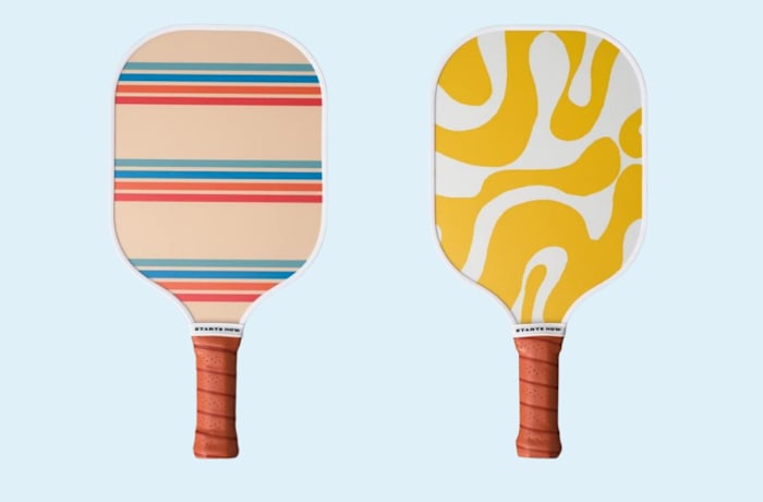 Two On Holiday pickleball paddles with orange handles, one hitting surface has a stripe design, and the other has a yellow and white gradient design.