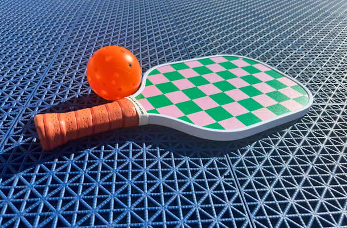 On Holiday pink and green checkered pickleball paddle with an orange handle on a blue textured surface next to an orange ball.