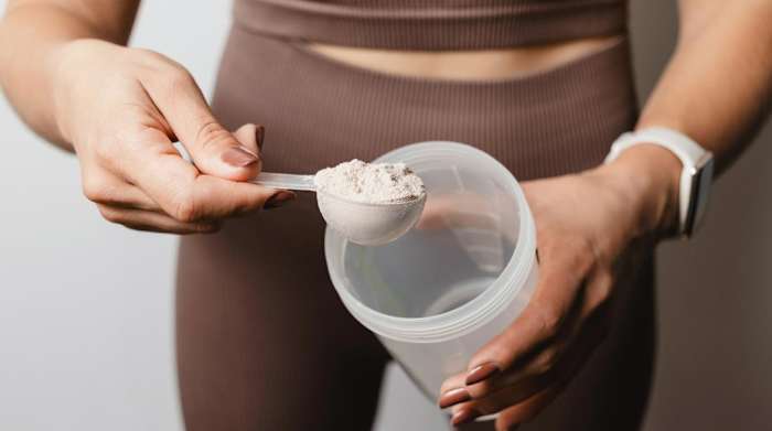 A woman wearing brown athletic clothes holding a shaker bottle and a scoop of creatine powder against a white background.