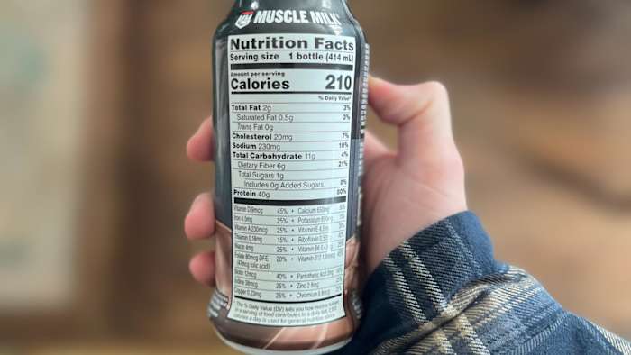 A hand holding a Muscle Milk Pro Advanced Nutrition Peanut Butter Chocolate protein shake with the nutrition label in view