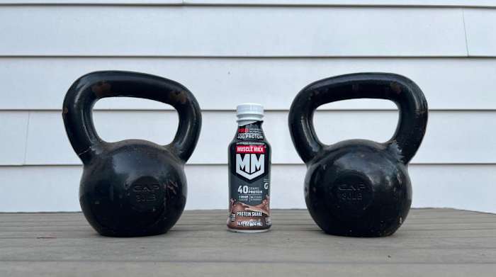 A white background with a table displaying a bottle of Muscle Milk Pro Advanced Nutrition Protein Shake between two kettlebells