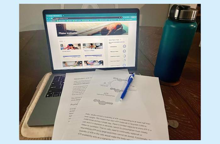 Study setup for ISSA Pilates Instructor course, with a lesson on a laptop, printouts for studying and a pen with a blue water bottle to the right of the computer