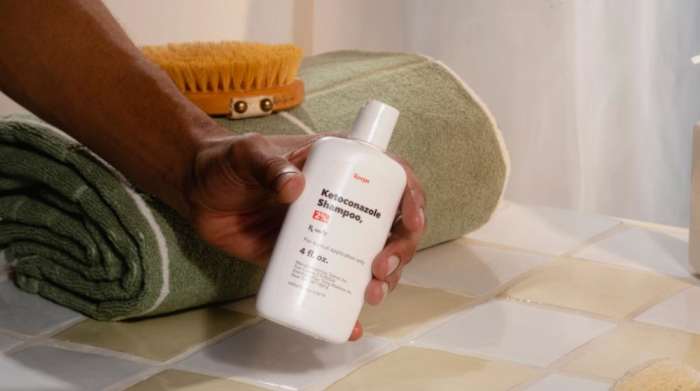 A man in a bathroom holding a bottle of Keeps ketoconazole shampoo for hair growth.