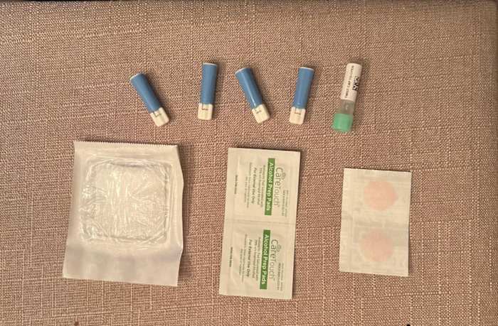 A brown background with four lancets, one gauze strip, one blood sample collection tube, two alcohol wipes and two bandages