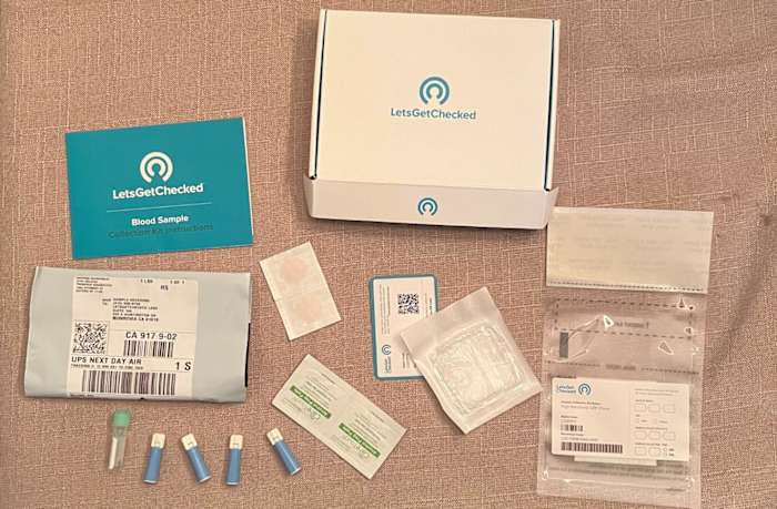 A brown background with a LetsGetChecked Heart Health kit laid out displaying the medical supplies (lancets, gauze, bandages, alcohol wipes), return shipping and collection labels