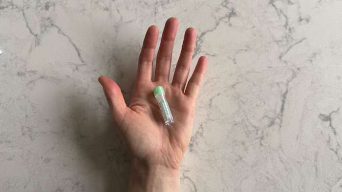 A woman holding a small vial for the LetsGetChecked Cholesterol test blood sample collection against a white and grey marble background.