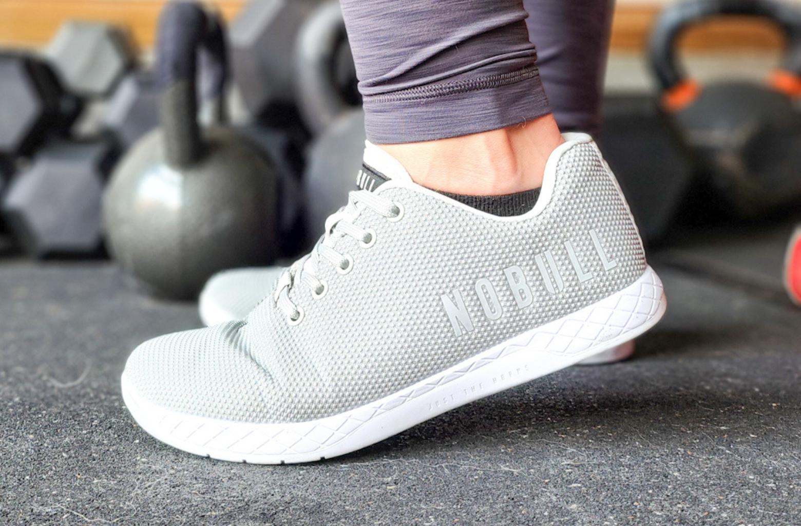 nobull-trainer-gray-on-feet-gray-leggings-black-socks-black-gym-floor-kettlebells-dumbeels-bars-in-background